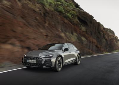 Audi A6 allroad (pearl grey) in a dynamic driving shot along a rocky coastal road.