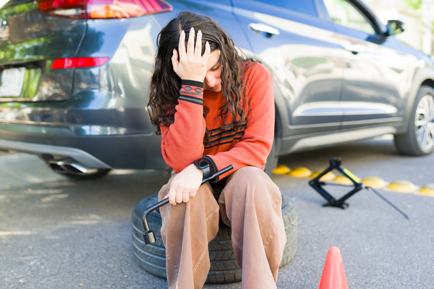 Teenage girl sitting on spare tire holding lug wrench with broken-down car and warning cone, experiencing frustration and stress