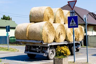 Modern agricultural tractor towing a trailer loaded with round hay bales through a rural village. Farming scene illustrating harvest logistics, agricultural productivity, sustainable farming, livestock feed production, and the daily operations of the rural economy.