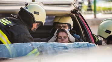 Close up shot through cut windshield of two firefighters helping injured female driver in shock, one is holding her head.